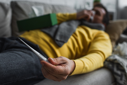 selective focus of electronic thermometer in hand of sick man lying on couch at home