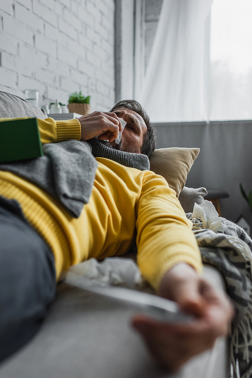 diseased man sneezing in paper napkin while lying on couch on blurred foreground