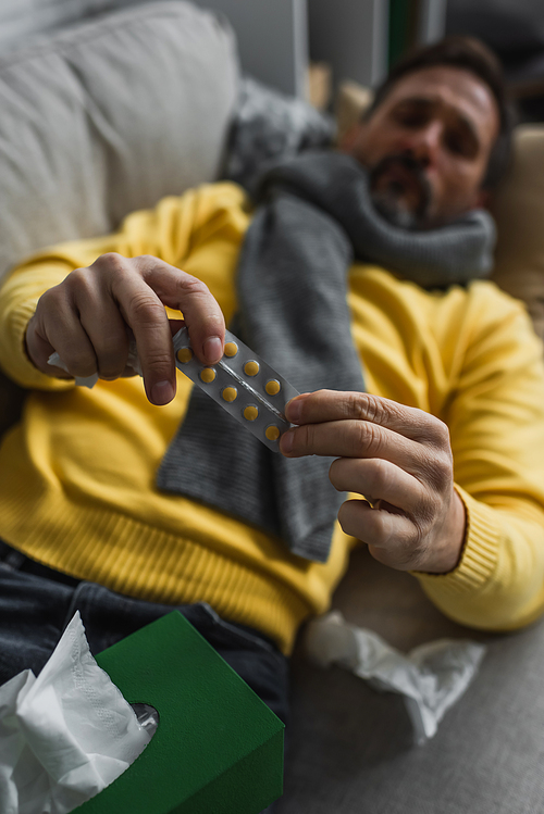 diseased man in warm scarf holding pills while lying on couch on blurred background