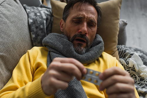 ill man in warm scarf holding blurred pills while lying on sofa