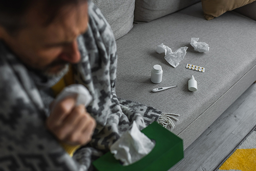 ill man with paper napkins sitting on couch near electronic thermometer and containers  with treatment