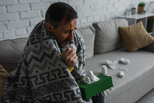 sick man holding pack of paper napkins while sitting on sofa near containers with treatment