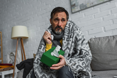 ill man sitting on couch under warm blanket and holding pack of paper napkins