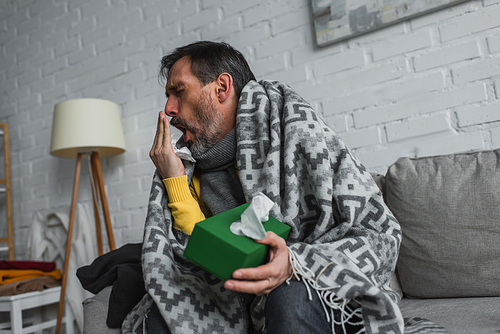 man covered with warm blanket holding pack of paper napkins and coughing on sofa