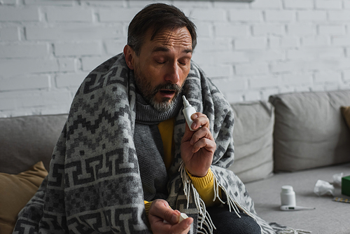 ill man with closed eyes sitting under warm blanket and holding nasal spray