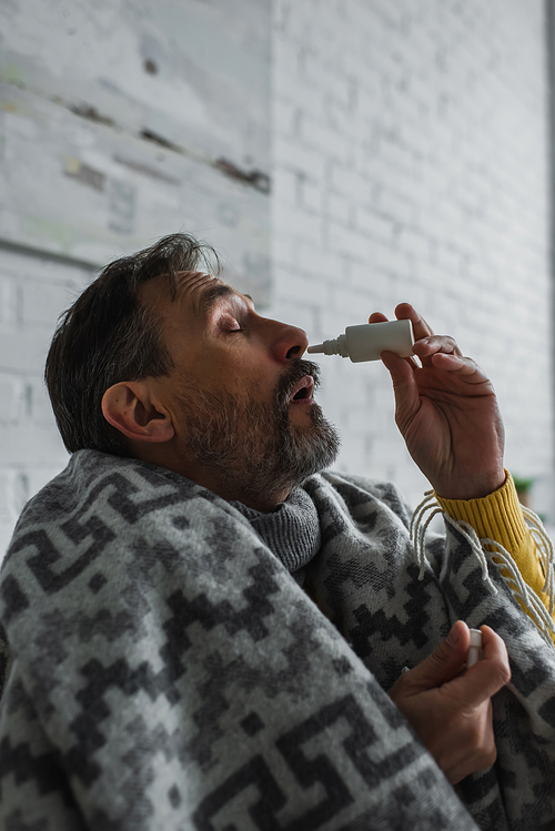 ill man suffering from runny nose and using nasal spray while sitting under warm blanket