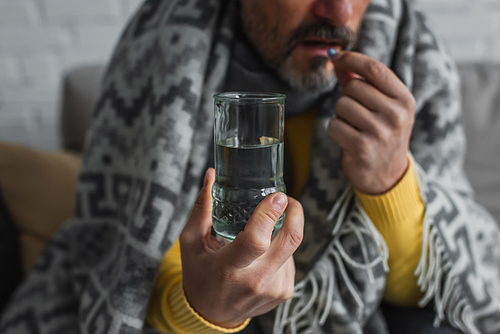 cropped view of sick man under warm blanket holding glass of water and taking pill on blurred background