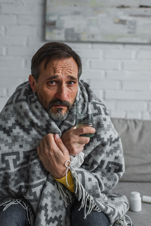 sick man sitting under warm blanket with glass of water and looking at camera