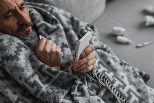 high angle view of sick man wrapped in warm blanket looking at mobile phone