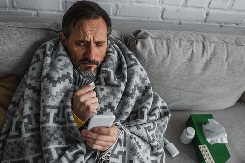 man covered with warm blanket looking at smartphone while sitting near containers with treatment and pack of paper napkins