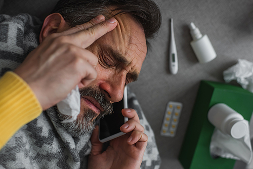 top view of sick man suffering from fever and headache while calling on smartphone near pills and containers with treatment