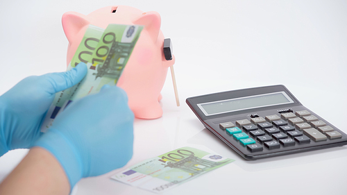cropped view of person in latex gloves counting euro banknotes near calculator and piggy bank on white background