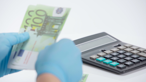 cropped view of person in latex gloves counting euro banknotes near calculator on white background