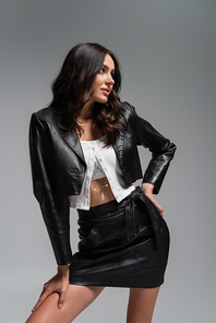 young woman in stylish black leather jacket and skirt posing with hand on hip isolated on grey