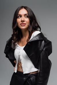 brunette young woman in black leather jacket looking away while posing isolated on grey