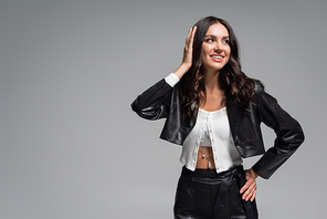 brunette young woman in stylish black leather jacket smiling and adjusting hair isolated on grey