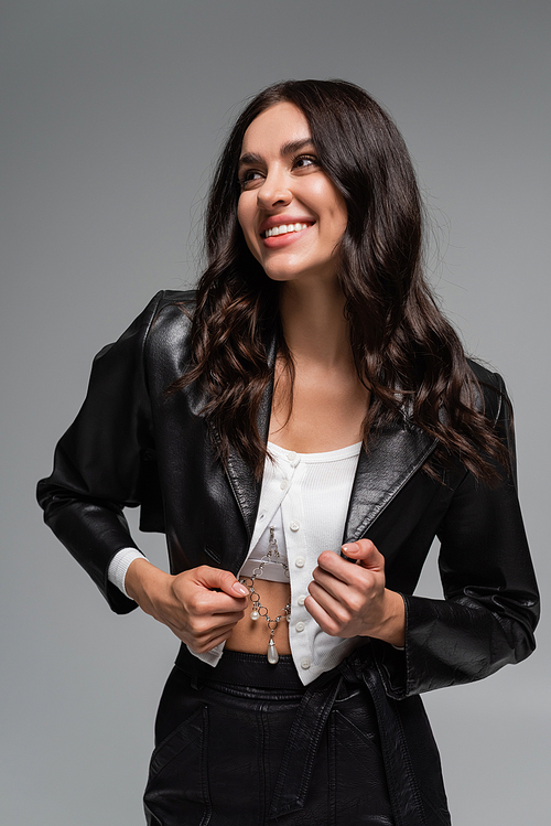 happy young woman adjusting black leather jacket isolated on grey