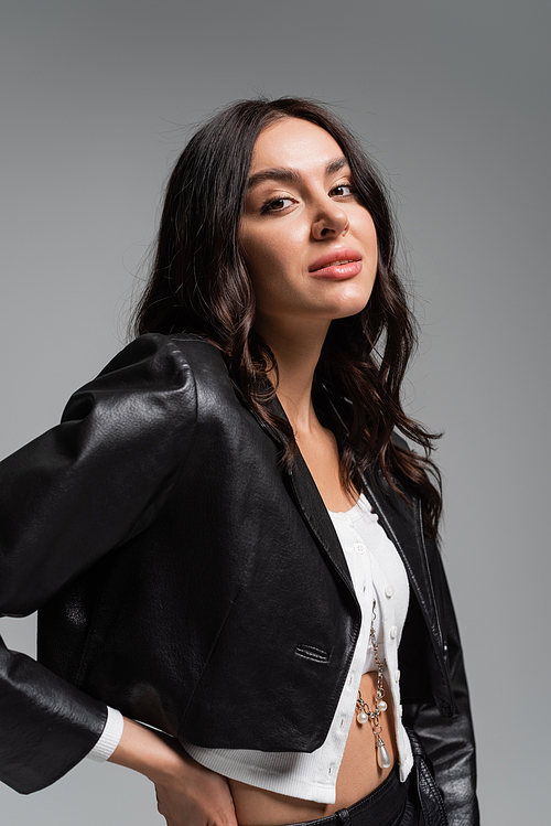 brunette young woman in stylish black leather jacket standing with hand on hip and looking at camera isolated on grey