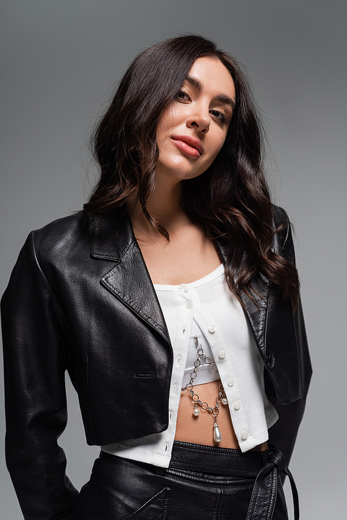 brunette young woman in stylish leather jacket looking at camera isolated on grey