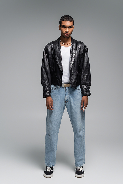 full length of young african american man in jeans and leather jacket on grey