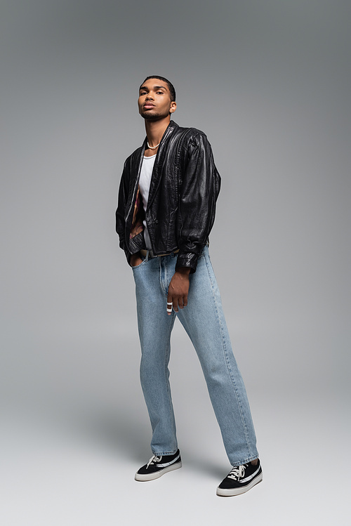 full length of young african american man in jeans and leather black jacket on grey