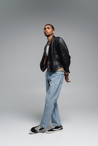 full length of young african american man in jeans and leather black jacket posing on grey