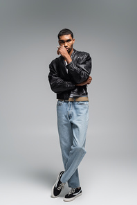 full length of stylish african american man in jeans and leather jacket posing on grey