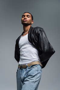 low angle view of young african american man in jeans and leather jacket posing isolated on grey