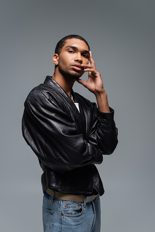young african american man in leather jacket touching face while posing isolated on grey