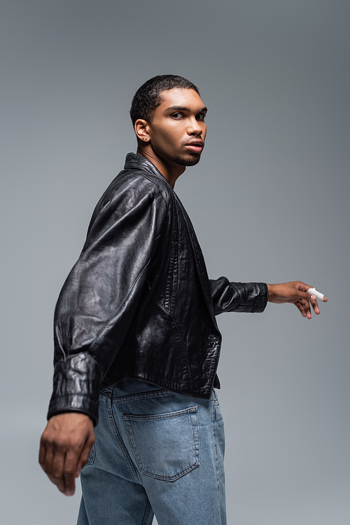young african american man in jeans and black leather jacket looking at camera isolated on grey