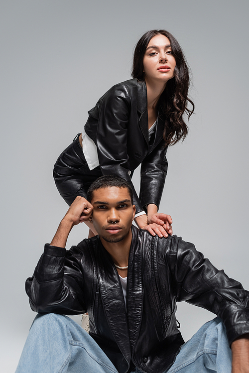stylish woman in leather jacket posing with young african american man isolated on grey