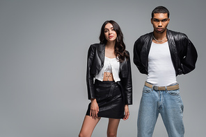 stylish woman in leather outfit posing with young african american man isolated on grey