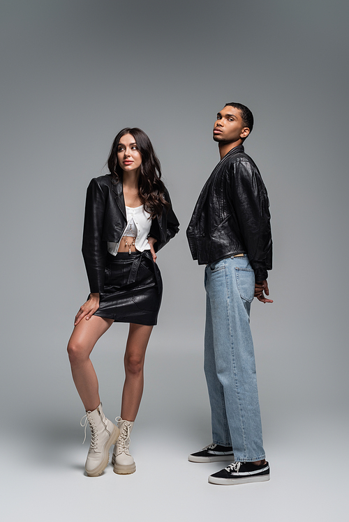 full length of stylish woman in leather outfit and boots posing with young african american man on grey