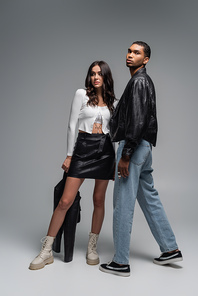 full length of brunette woman in leather skirt holding black jacket near stylish african american man on grey
