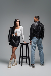 full length of woman in trendy outfit standing near high chair and stylish african american man on grey