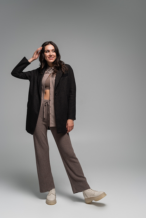 full length of cheerful young woman in black blazer and knitted trousers posing on grey