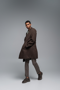 full length of young african american man in autumnal woolen coat looking at camera and posing with hand in pocket on grey