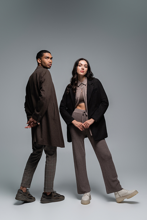 full length of stylish interracial models in trendy autumnal outfits posing together on grey