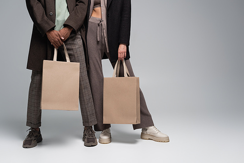 cropped view of stylish interracial couple in autumnal outfits holding shopping bags on grey