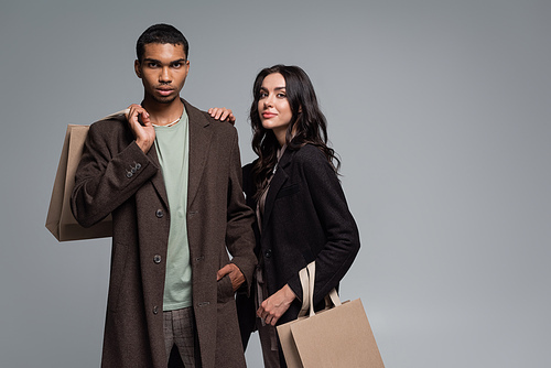 stylish interracial couple in autumnal outfits holding shopping bags isolated on grey