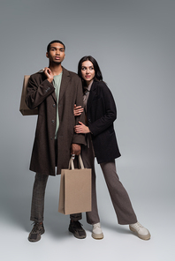 full length of stylish interracial couple in trendy autumnal outfits holding shopping bags on grey