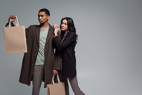 stylish woman in black blazer near young african american man in coat holding shopping bags isolated on grey