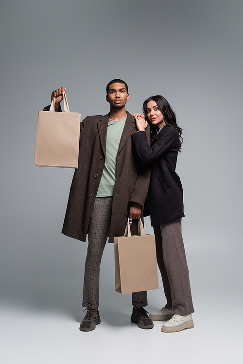 full length of stylish interracial couple in autumnal outfits holding paper bags on grey