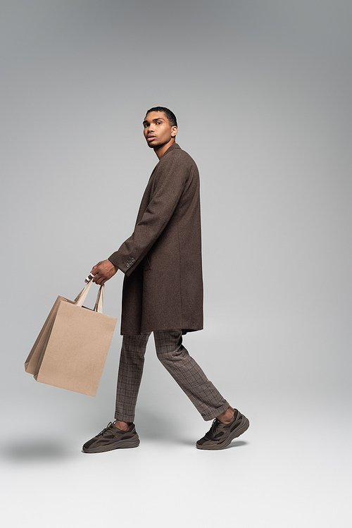 full length of stylish african american man in autumnal coat and sneakers walking with shopping bags on grey