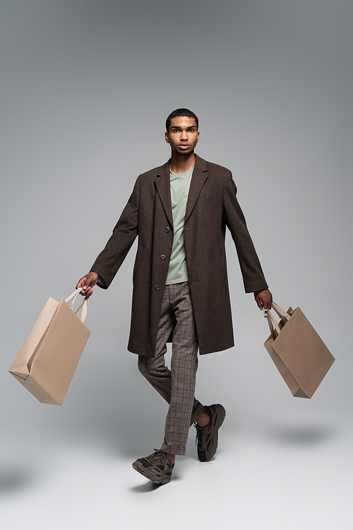 full length of stylish african american man in woolen coat and sneakers posing with shopping bags on grey