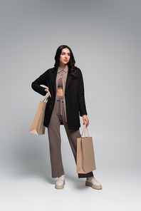 full length of young woman in stylish autumnal outfit standing with shopping bags on grey
