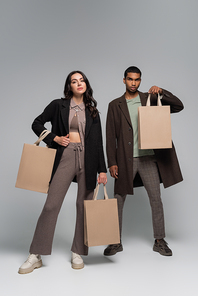 full length of stylish interracial models in autumnal outfits holding shopping bags on grey