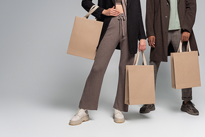 partial view of stylish interracial couple in autumnal outfits holding shopping bags on grey