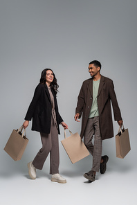 full length of happy interracial couple in stylish outfits holding shopping bags and walking on grey