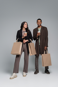 full length of happy interracial couple in stylish outfits holding shopping bags and paper cups on grey